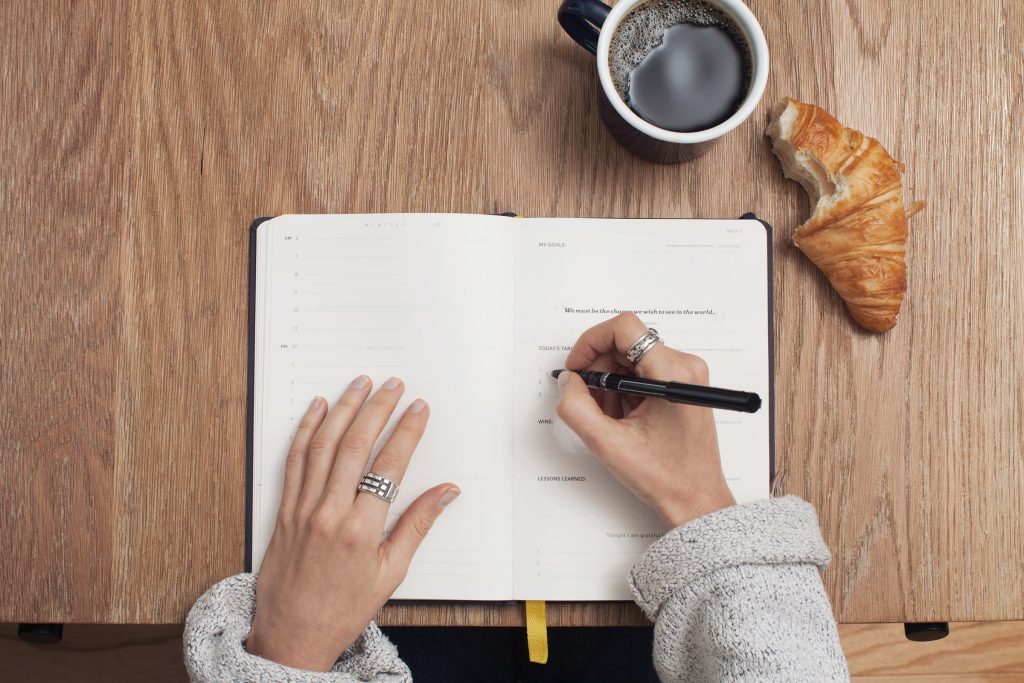 femme écrivant ses tâches sur son agenda à côté d'une tasse à café et un croissant